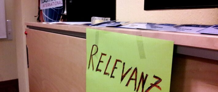 Rock’n Roll ist mehr als nur Musik Handgeschriebenes grünes Schild mit dem Wort „Relevanz“ liegt auf einem Tisch vor dem Toastmasters-Logo.