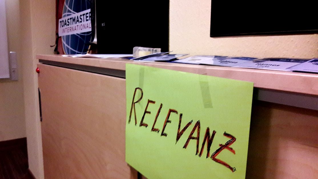 Handgeschriebenes grünes Schild mit dem Wort „Relevanz“ liegt auf einem Tisch vor dem Toastmasters-Logo.