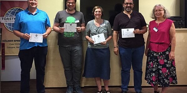 Die ultimative Röstparty Fünf lächelnde Mitglieder der Kölner Sprechbar stehen vor dem Toastmasters-Banner und halten ihre Urkunden in den Händen.