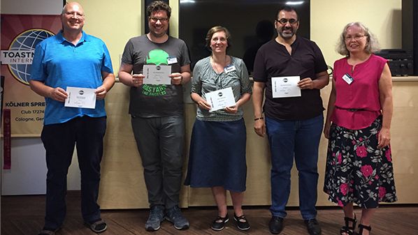 Fünf lächelnde Mitglieder der Kölner Sprechbar stehen vor dem Toastmasters-Banner und halten ihre Urkunden in den Händen.