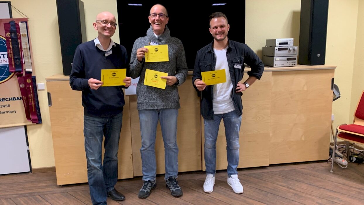 Drei lächelnde Toastmasters-Mitglieder mit ihren gelben Auszeichnungsurkunden vor einem Vereinsbanner.