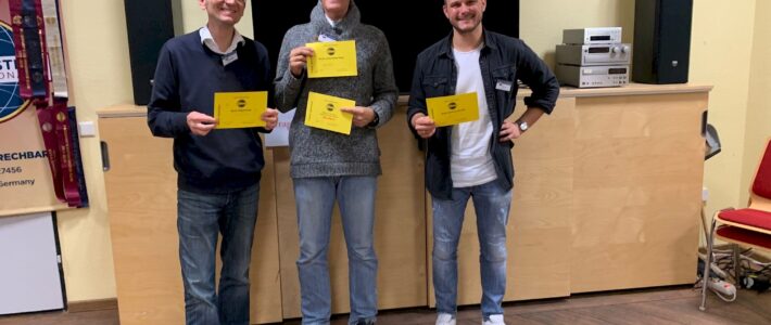 Drei lächelnde Toastmasters-Mitglieder mit ihren gelben Auszeichnungsurkunden vor einem Vereinsbanner.