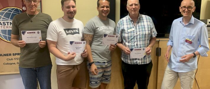 Sommer, Sonne, Sockenlöcher! Fünf Teilnehmer der Kölner Sprechbar stehen bei einem Clubabend mit Urkunden vor Toastmasters-Banner und Bildschirm.