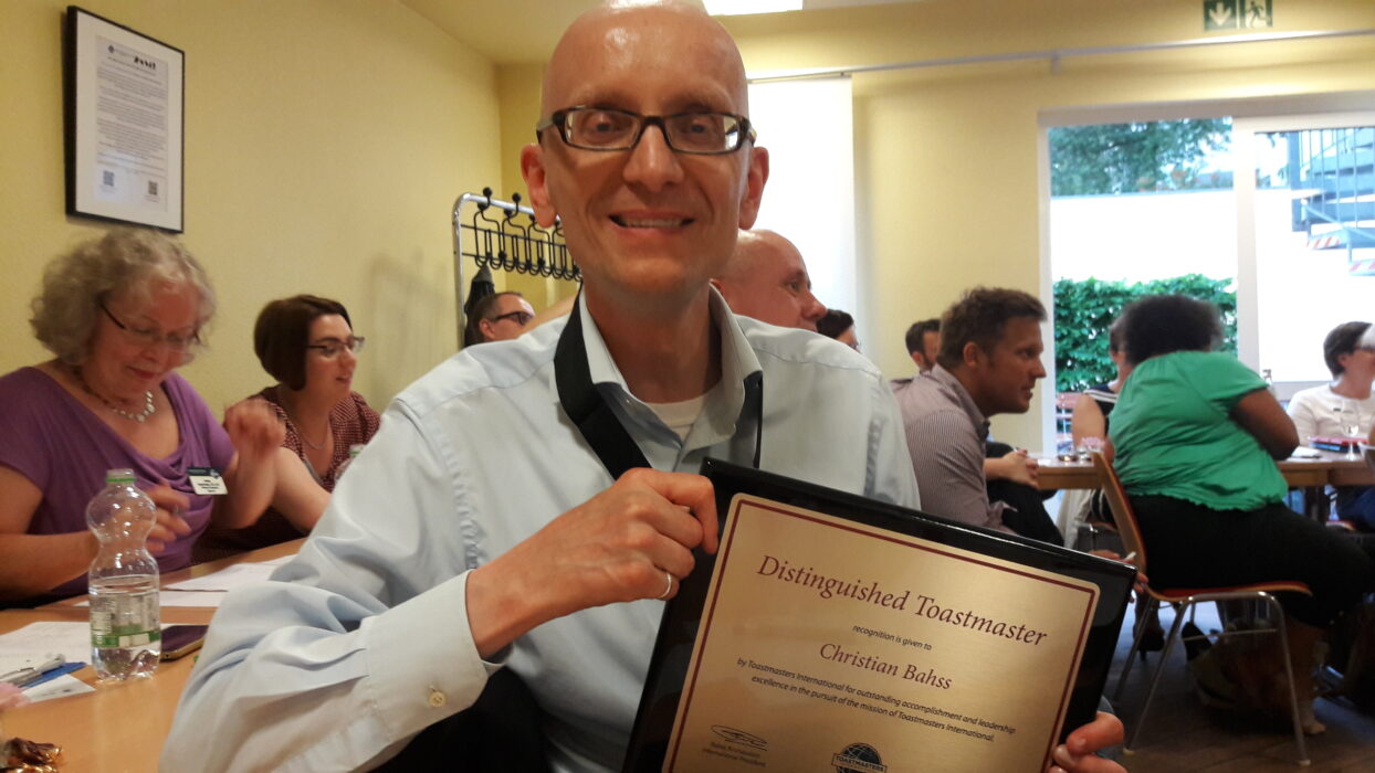 Christian Bahß hält lächelnd seine Urkunde als Distinguished Toastmaster in die Kamera, im Hintergrund weitere Mitglieder der Kölner Sprechbar.