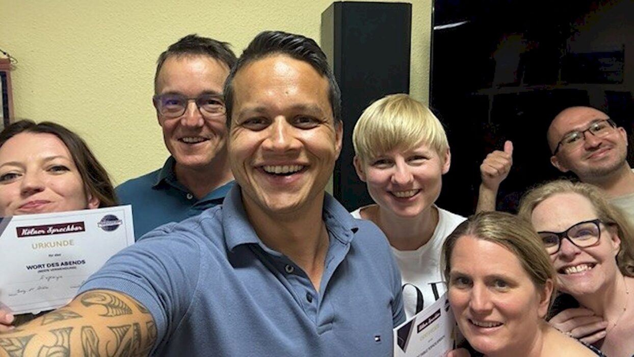 Seven smiling Kölner Sprechbar members pose for a group selfie after the meeting, several holding award certificates and one giving a thumbs-up, in front of a light-coloured wall with a loudspeaker in the background.
