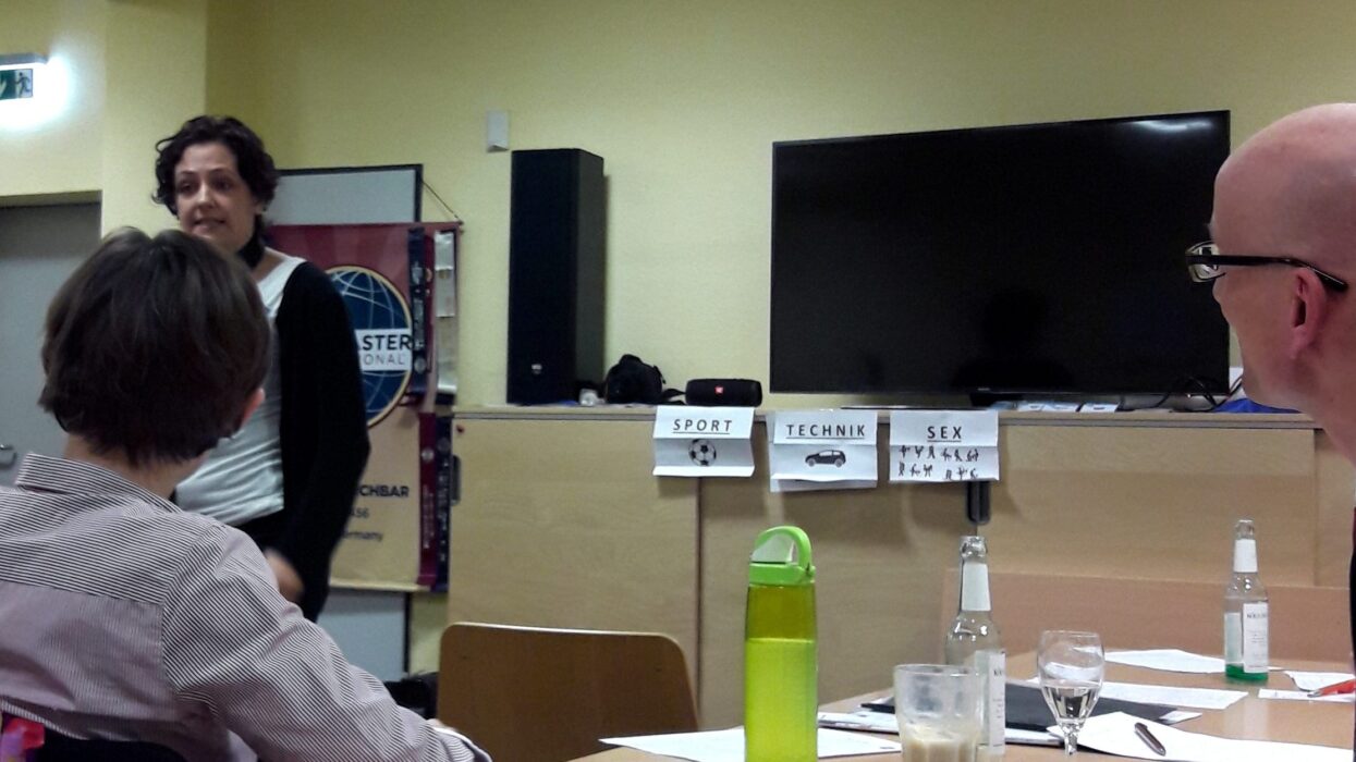 Eine Toastmasterin präsentiert vor dem Publikum der Kölner Sprechbar, im Hintergrund Kärtchen mit den Themen Sport, Technik und Sex.