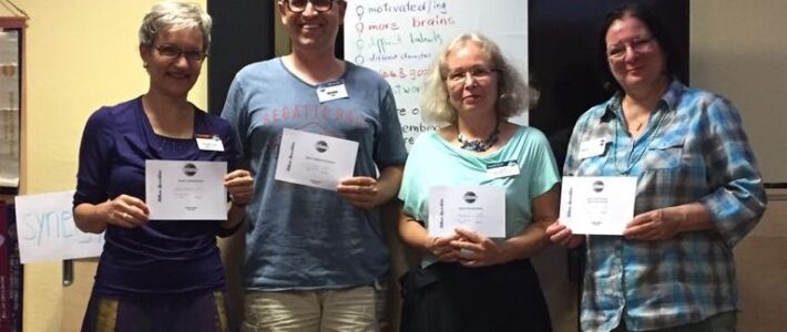 Vier lächelnde Mitglieder der Kölner Sprechbar stehen vor einem Flipchart mit der Aufschrift „A good team is“ und halten ihre Urkunden in den Händen.