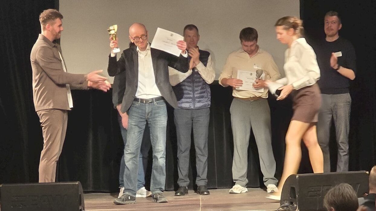 Christian B. hält bei der Siegerehrung des Toastmasters Division-Wettbewerbs 2026 einen Pokal hoch, weitere Ausgezeichnete auf der Bühne.