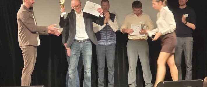 Congratulazioni fantastiche: Kölner Sprechbar erfolgreich auf Divisionsebene Christian B. hält bei der Siegerehrung des Toastmasters Division-Wettbewerbs 2026 einen Pokal hoch, weitere Ausgezeichnete auf der Bühne.