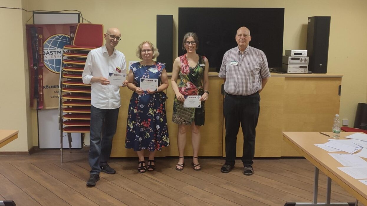 Vier Personen stehen nebeneinander in einem Seminarraum. Drei von ihnen halten Urkunden in den Händen. Im Hintergrund sind ein Toastmasters-Banner, gestapelte Stühle, ein großer schwarzer Bildschirm und eine Holztheke zu sehen.