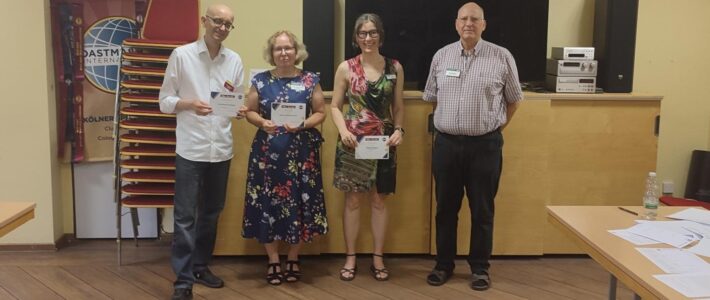 Finanzen sind Chefsache: Psychologische Fallen beim Investieren Vier Personen stehen nebeneinander in einem Seminarraum. Drei von ihnen halten Urkunden in den Händen. Im Hintergrund sind ein Toastmasters-Banner, gestapelte Stühle, ein großer schwarzer Bildschirm und eine Holztheke zu sehen.