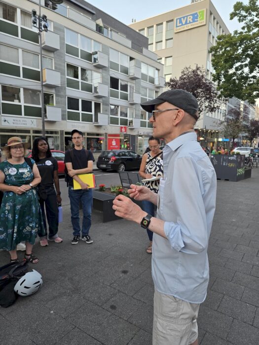 Ein Redner spricht auf einem Platz in Köln-Deutz, vier Zuhörende stehen ihm gegenüber, im Hintergrund ein Bürogebäude.