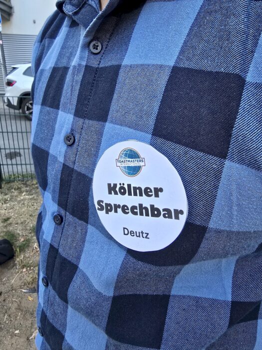 Nahaufnahme eines Ansteckers mit Toastmasters-Logo und der Aufschrift Kölner Sprechbar Deutz auf einem karierten Hemd.