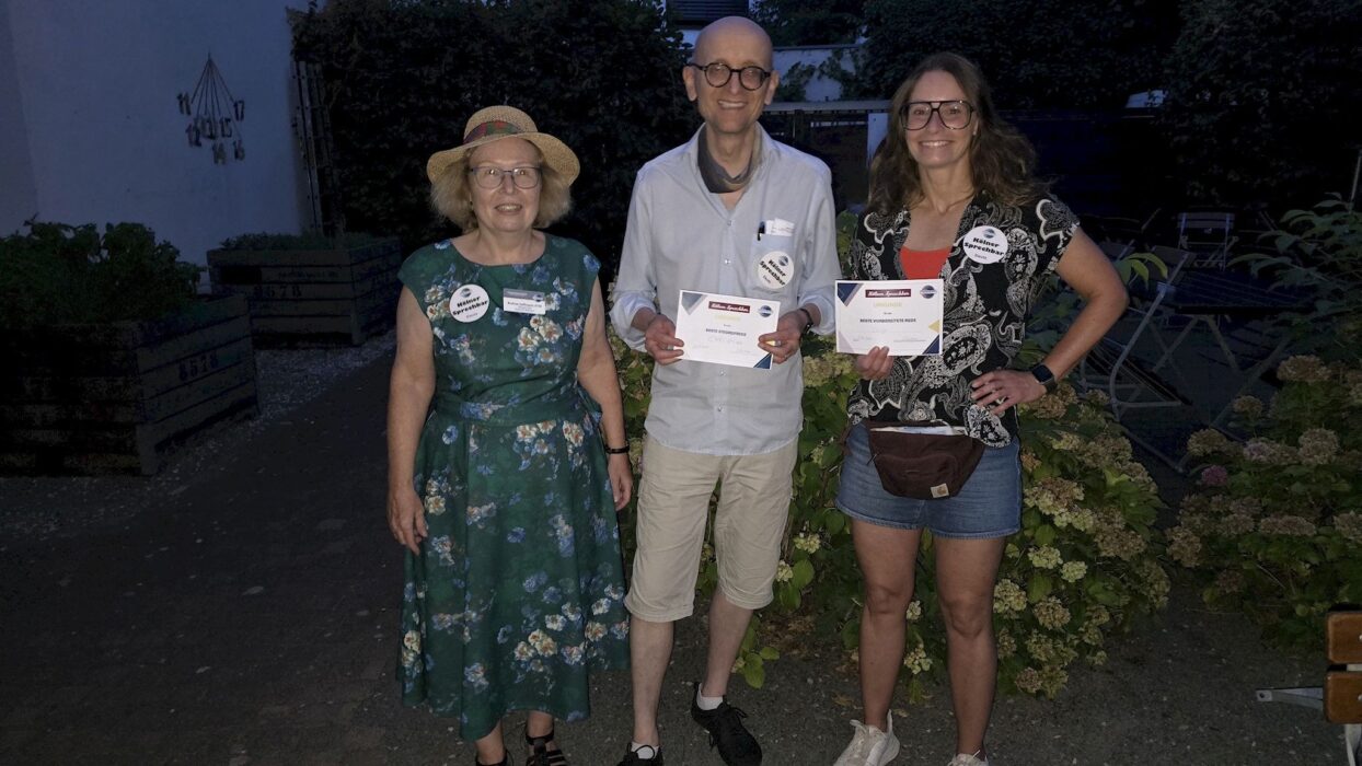 Drei Mitglieder der Kölner Sprechbar stehen abends im Innenhof des Bürgerzentrums, zwei von ihnen halten Urkunden für die beste Stegreifrede und die beste vorbereitete Rede.