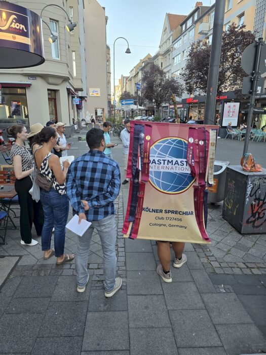 Eine Person trägt das Banner der Kölner Sprechbar durch eine Einkaufsstraße in Deutz, weitere Mitglieder gehen daneben.