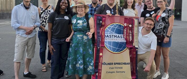 Rund vierzehn Mitglieder und Gäste der Kölner Sprechbar stehen bei einem Stadtrundgang in Köln-Deutz vor einem historischen Gebäude und halten das Clubbanner mit dem Toastmasters-Logo.