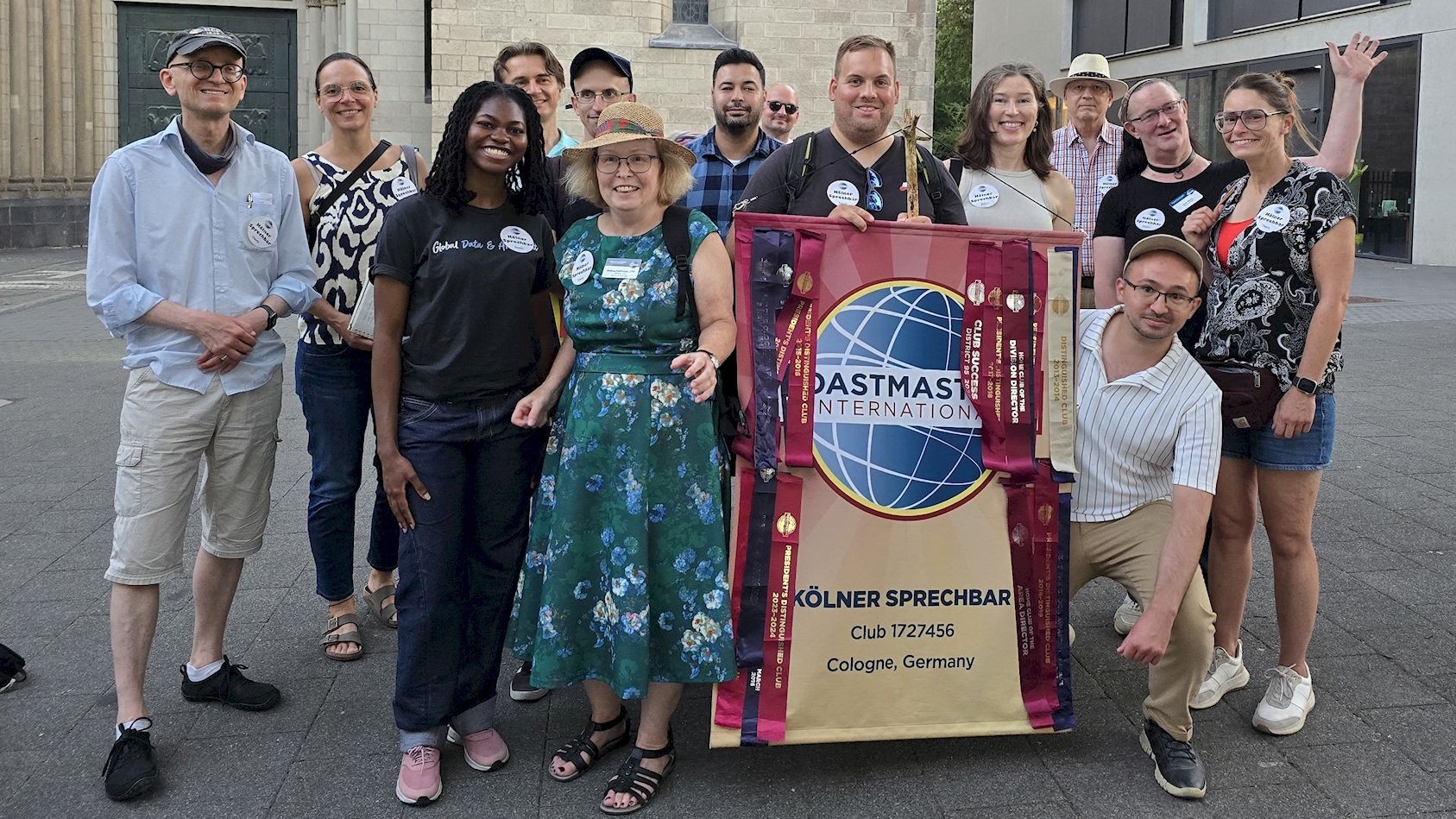 Rund vierzehn Mitglieder und Gäste der Kölner Sprechbar stehen bei einem Stadtrundgang in Köln-Deutz vor einem historischen Gebäude und halten das Clubbanner mit dem Toastmasters-Logo.