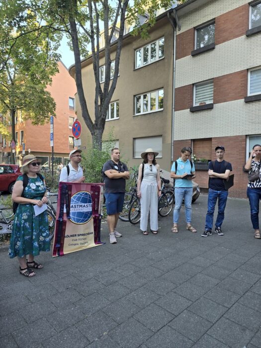 Mitglieder der Kölner Sprechbar stehen mit dem Clubbanner auf einem Gehweg in Köln-Deutz und hören zu.