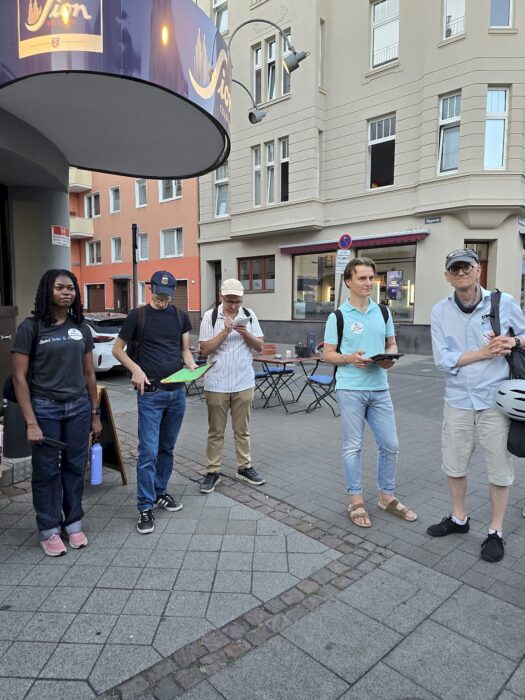 Fünf Mitglieder der Kölner Sprechbar stehen auf dem Gehweg unter einem Brauhausschild, eine Person macht Notizen.