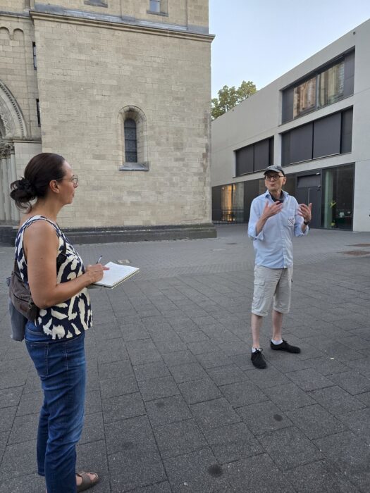 Ein Redner spricht mit erhobenen Händen auf einem Platz vor einer romanischen Kirche, eine Zuhörerin notiert mit.