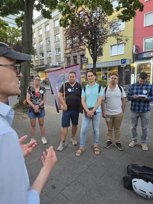 Ein Redner gestikuliert im Vordergrund, fünf Zuhörende stehen vor dem Clubbanner auf dem Gehweg.