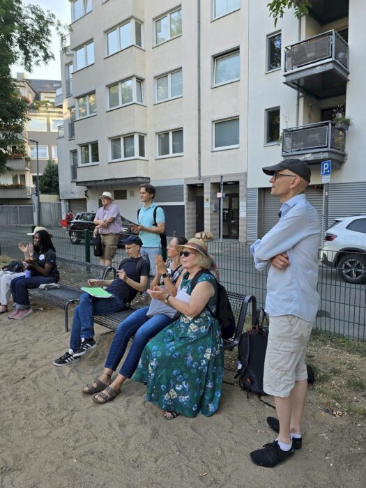 Zuhörende sitzen auf einer Bank am Spielplatz und applaudieren, weitere Personen stehen daneben.
