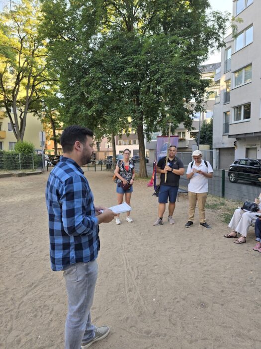 Ein Redner spricht mit Notizen in der Hand auf einem sandigen Spielplatz, im Hintergrund hält jemand das Clubbanner an einem Holzstock.