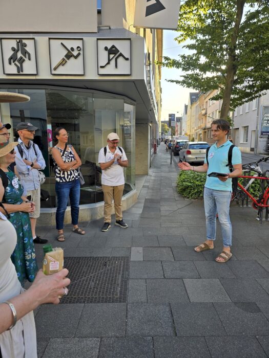Ein Redner spricht auf dem Gehweg vor einem Ladenlokal mit Sport-Piktogrammen, mehrere Zuhörende stehen im Halbkreis.