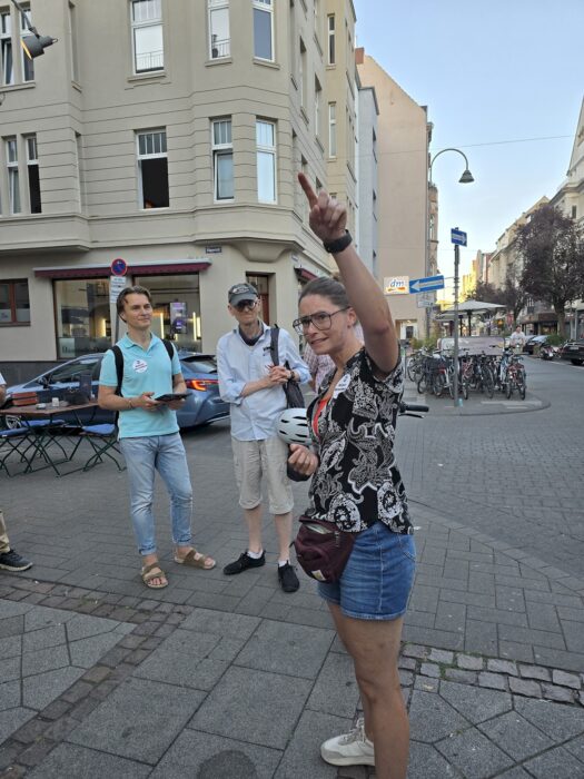 Eine Rednerin zeigt beim Sprechen nach oben, zwei Zuhörende stehen an einer Straßenecke daneben.