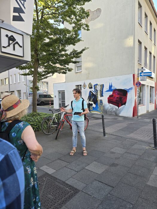 Ein Redner steht vor einer Wandmalerei mit Kölner Motiven wie Dom, Brücke und rotem Sessel und spricht zur Gruppe.