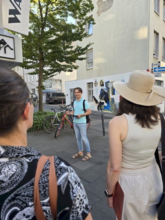 Ein Redner spricht an einer Straßenecke vor einer Wandmalerei, zwei Zuhörende stehen im Vordergrund.