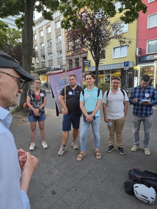 Fünf Zuhörende stehen vor dem Clubbanner, im Vordergrund ist der sprechende Redner angeschnitten zu sehen.