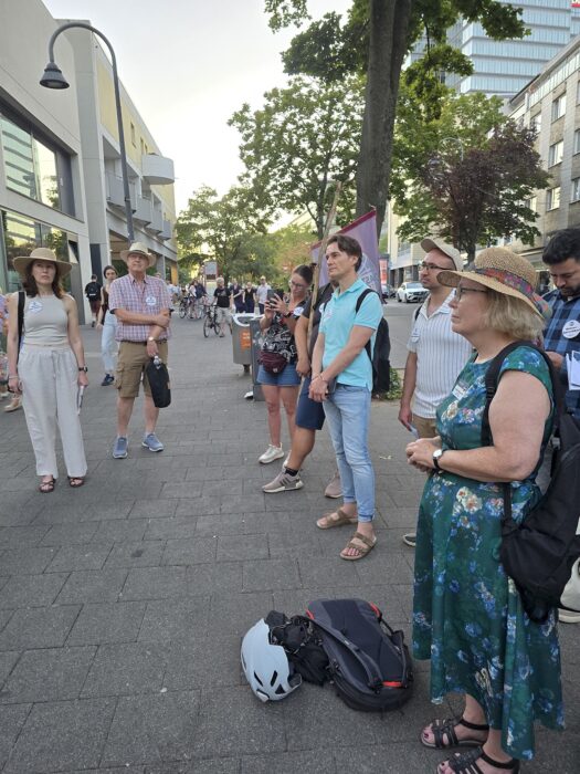 Mitglieder und Gäste der Kölner Sprechbar hören auf einem breiten Gehweg zu, das Clubbanner steht an einem Holzstock.