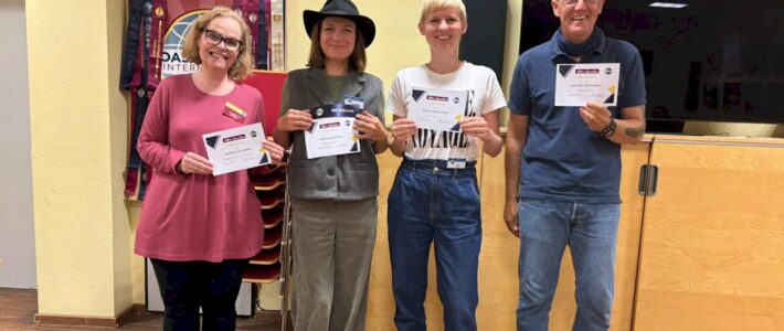 Four award recipients stand in the club room holding certificates; one wears a black hat and holds an Ice Breaker ribbon.