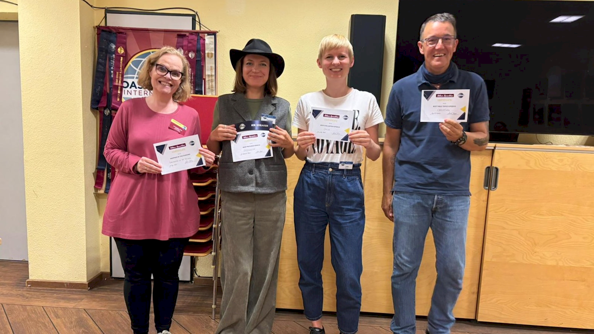 Four award recipients stand in the club room holding certificates; one wears a black hat and holds an Ice Breaker ribbon.