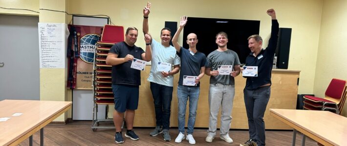 Fünf Personen stehen lächelnd nebeneinander im Clubraum und halten Urkunden. Mehrere heben einen Arm. Im Hintergrund sind ein Toastmasters-Banner, gestapelte Stühle und ein großer Bildschirm zu sehen.