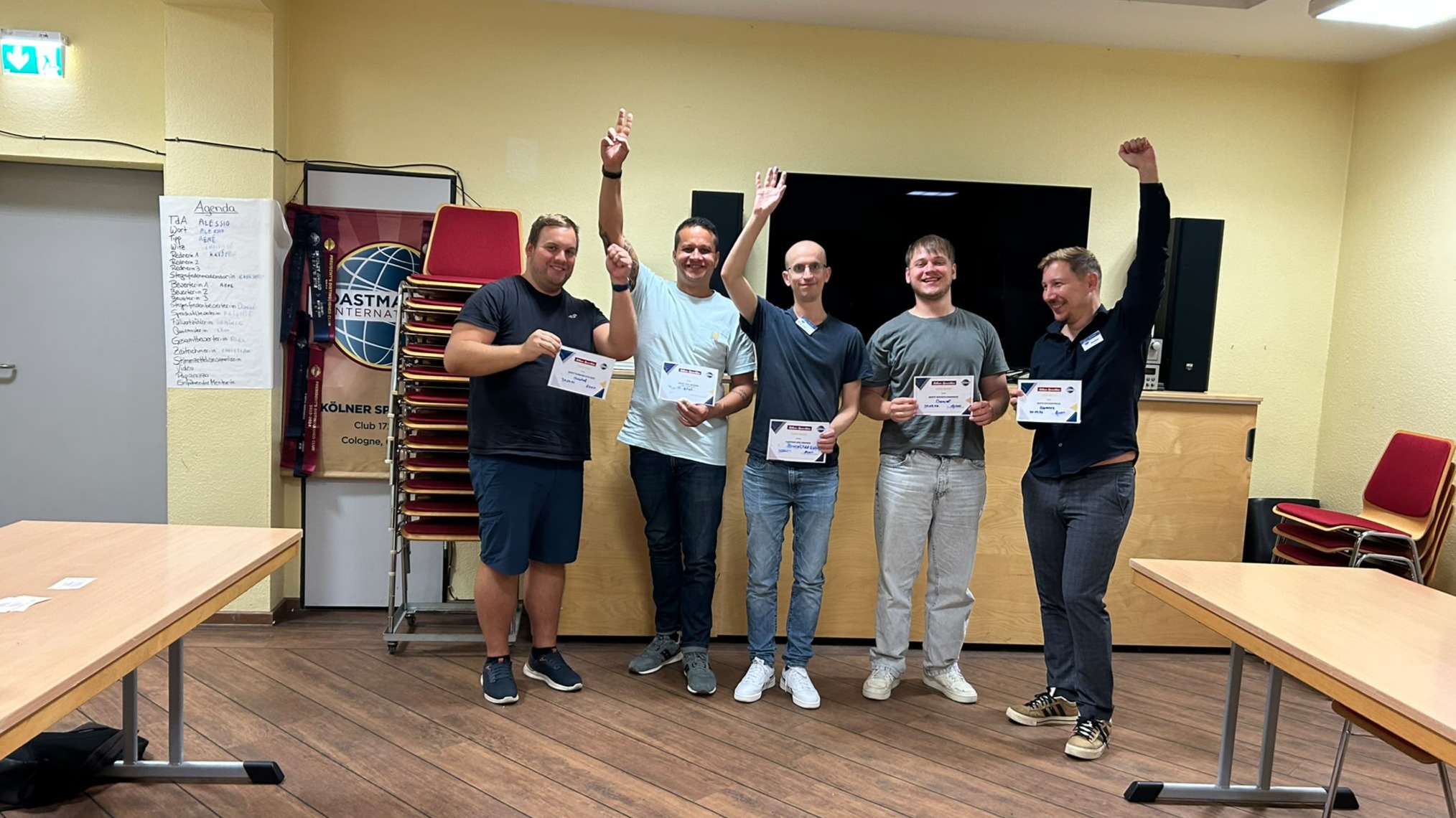 Fünf Personen stehen lächelnd nebeneinander im Clubraum und halten Urkunden. Mehrere heben einen Arm. Im Hintergrund sind ein Toastmasters-Banner, gestapelte Stühle und ein großer Bildschirm zu sehen.