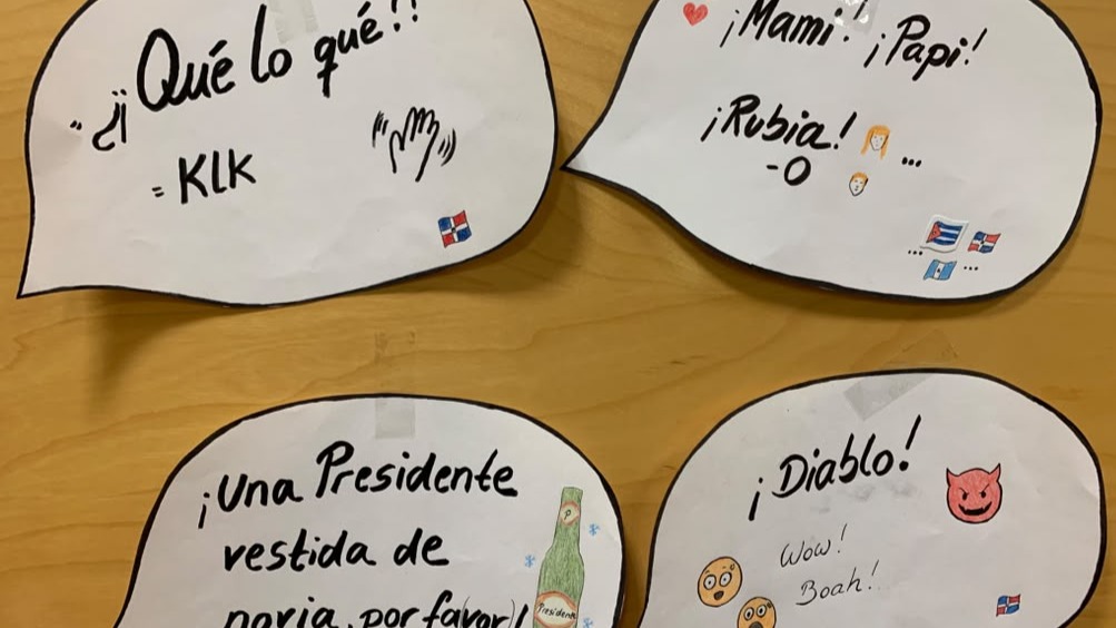 Vier weiße Sprechblasen auf einem Holztisch mit spanischen Ausrufen wie „¡Qué lo qué?!“, „¡Mami! ¡Papi!“, „¡Una Presidente vestida de novia, por favor!“ und „¡Diablo!“, dazu kleine Flaggen und Symbole.