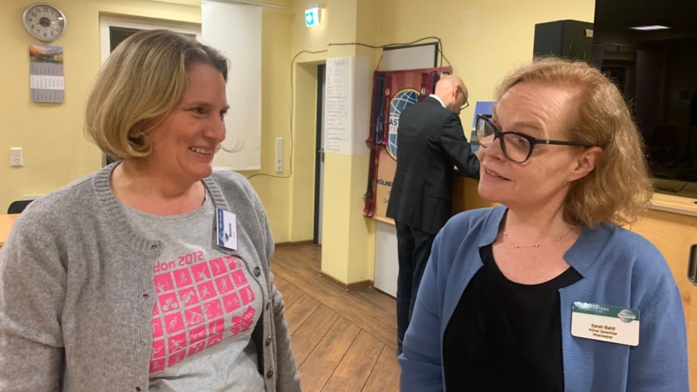 Zwei Frauen unterhalten sich lächelnd in einem Raum, eine im grauen Cardigan, eine in blauer Strickjacke mit Brille. Im Hintergrund steht ein Mann an einem Pult vor einem Banner.