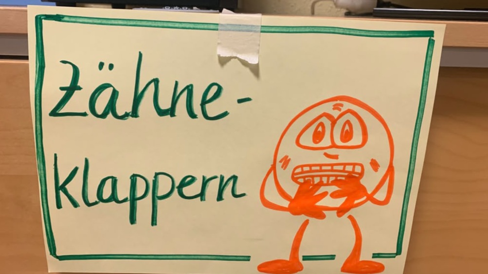 Handgezeichnetes Schild mit dem Text „Zähne-Klappern“ in grüner Schrift und einer orangefarbenen Comicfigur mit weit aufgerissenen Augen und klappernden Zähnen.