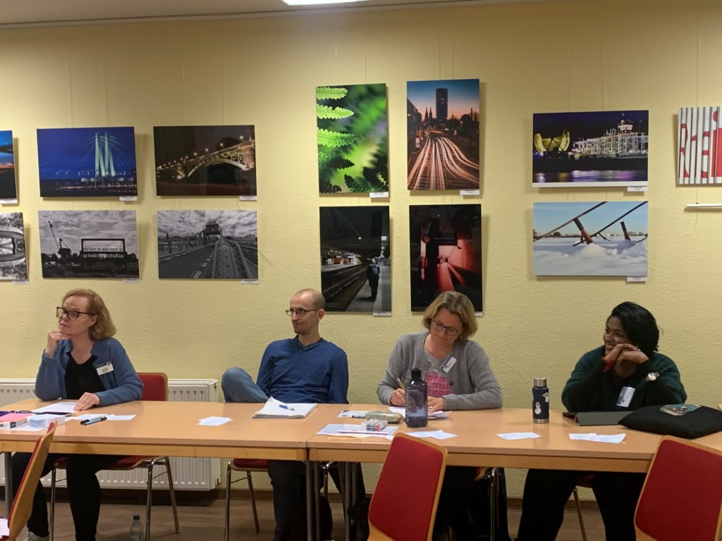 Vier Personen sitzen an Tischen vor einer Wand mit großformatigen Fotografien, machen sich Notizen oder hören zu.