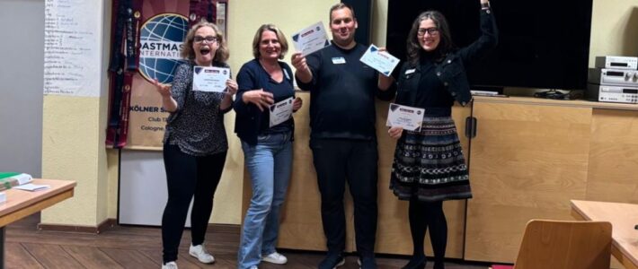 Four people stand together in a club room holding certificates. The person on the right raises an arm. A Toastmasters banner, stacked chairs and a large screen are visible behind them.