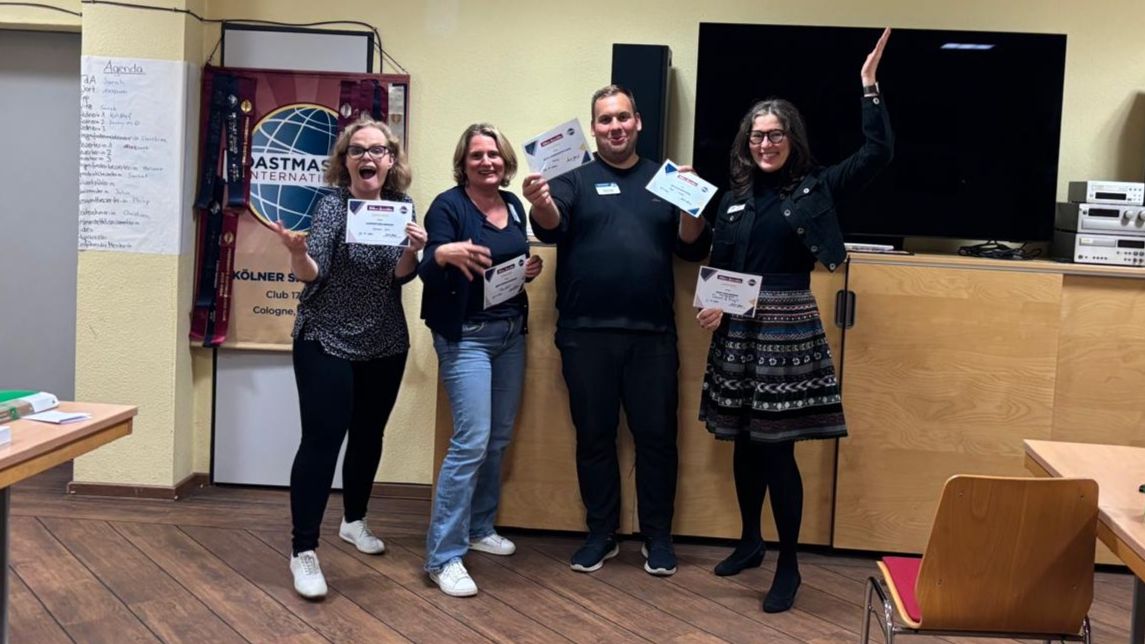 Four people stand together in a club room holding certificates. The person on the right raises an arm. A Toastmasters banner, stacked chairs and a large screen are visible behind them.