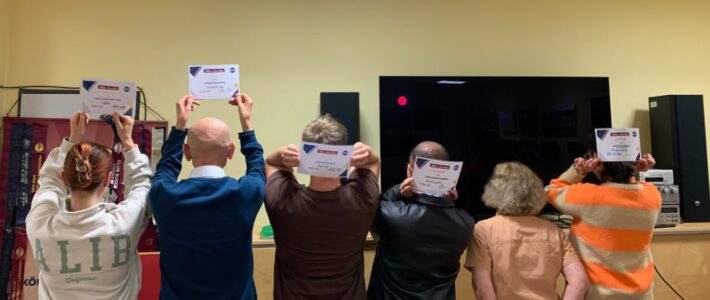Sechs Personen stehen mit dem Rücken zur Kamera im Clubraum. Fünf halten Urkunden über dem Kopf oder hinter dem Kopf. Im Hintergrund stehen ein Toastmasters-Banner und ein großer Bildschirm.