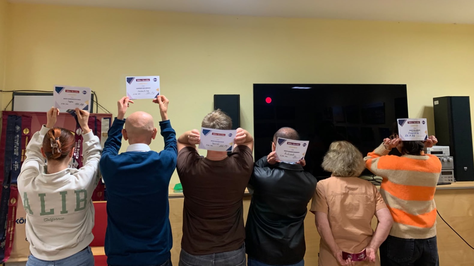 Sechs Personen stehen mit dem Rücken zur Kamera im Clubraum. Fünf halten Urkunden über dem Kopf oder hinter dem Kopf. Im Hintergrund stehen ein Toastmasters-Banner und ein großer Bildschirm.